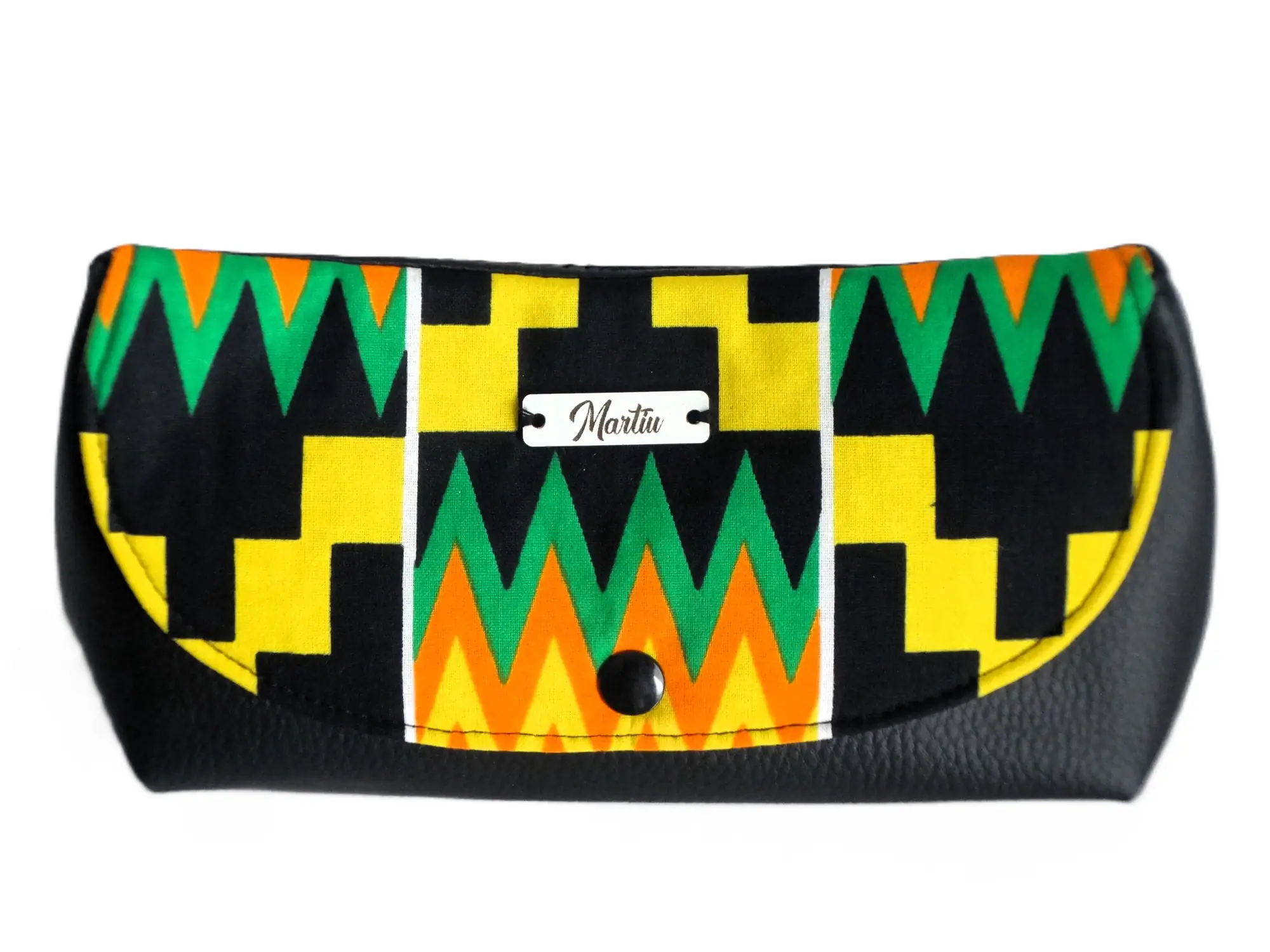 Cartera de mano de la colección de MARTIU que combina una base de cuero negro con una solapa de tela artesanal de patrones vibrantes en zig-zag verdes y naranjas, incluyendo el logotipo central de la marca.