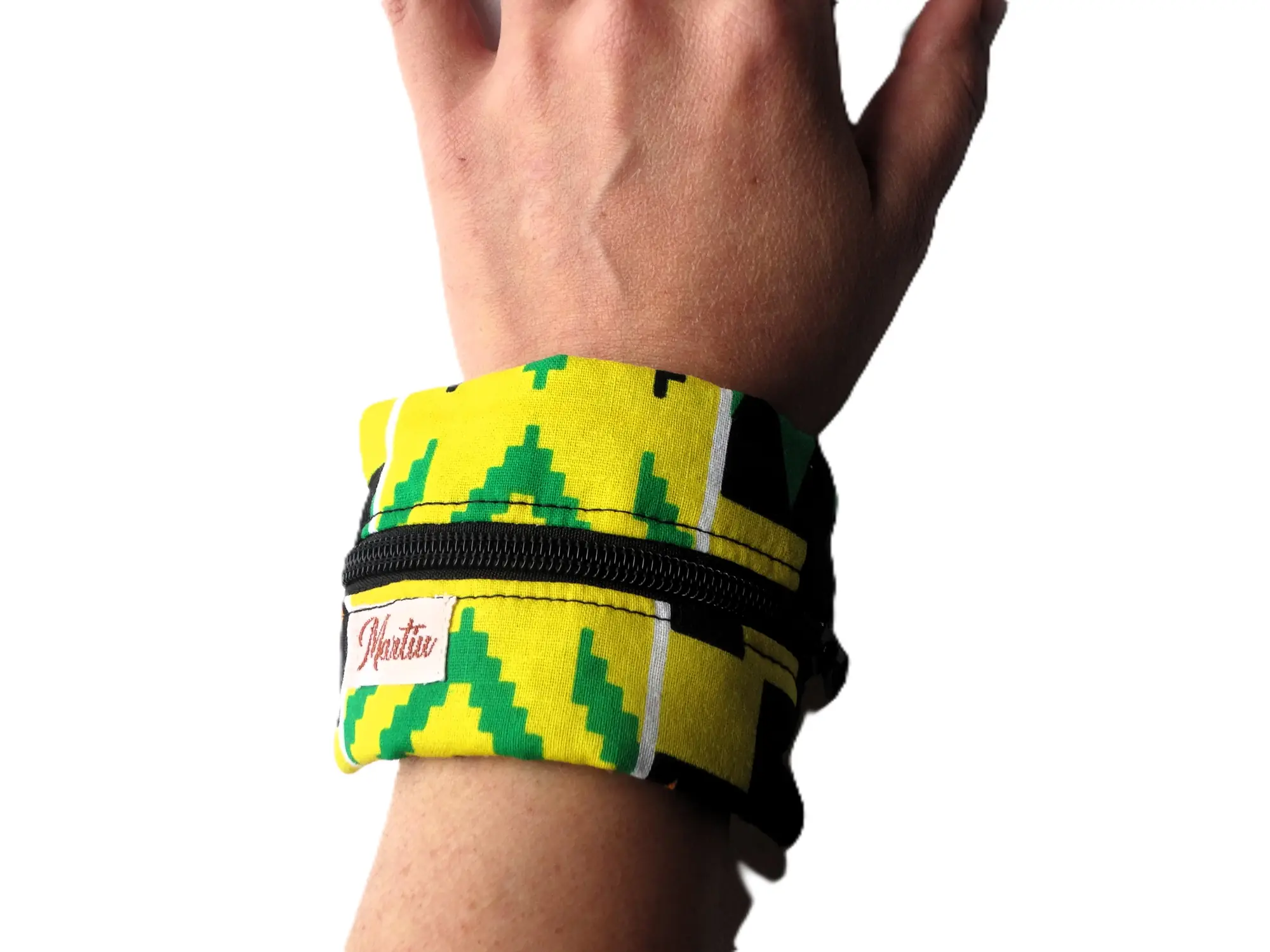 Detalle de una pulsera monedero de MARTIU colocada en la muñeca, fabricada con tela de colores amarillo y verde con cierre de cremallera negro, un accesorio innovador que integra la etiqueta original de la marca.
