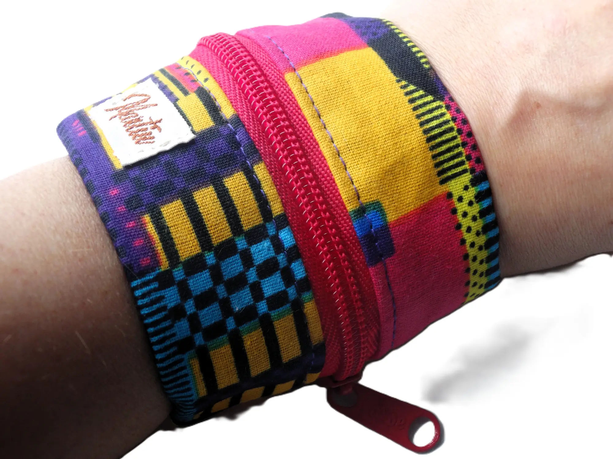 Detalle de una pulsera monedero de la marca MARTIU colocada en la muñeca, que muestra un patrón textil vibrante en rosa, amarillo y azul con una cremallera roja para guardar pequeños objetos de forma segura.